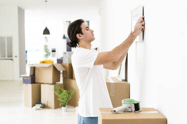 Young man putting up picture frame on wall at his new apartment - GIOF09727