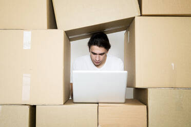 Young man working on laptop under stack of cardboard boxes - GIOF09731