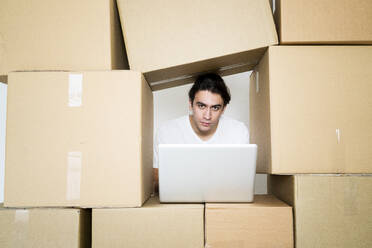Young man with laptop while trapped between cardboard boxes at new apartment - GIOF09732