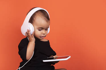 Niedliches kleines Mädchen mit Kopfhörern, das ein Mobiltelefon benutzt, während es vor einem orangefarbenen Hintergrund sitzt - RTBF01493