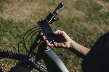 Frau, die ein Mobiltelefon benutzt, während sie in einem Park neben einem elektrischen Mountainbike steht - DMGF00304