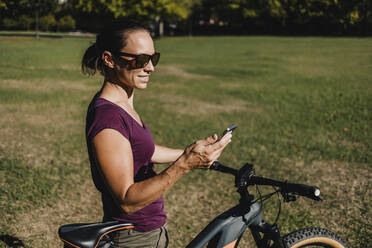 Smiling female athlete using mobile phone while standing with electric mountain bike at park - DMGF00305
