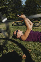 Sportswoman using mobile phone while resting on grass by electric mountain bike at park - DMGF00307