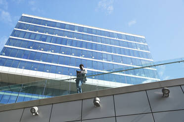 Geschäftsmann, der ein Smartphone benutzt, während er am Glasgeländer eines Bürogebäudes steht - PMF01545