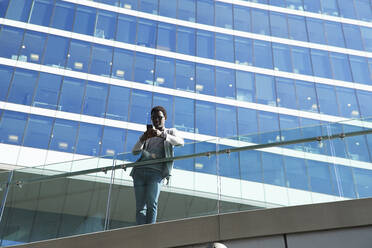 Männlicher Berufstätiger, der ein Smartphone benutzt, während er an einem Glasgeländer vor einem Bürogebäude steht - PMF01546