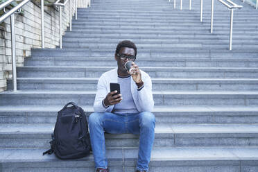 Geschäftsmann trinkt Kaffee und sitzt mit seinem Smartphone auf einer Treppe im Stadtzentrum - PMF01550