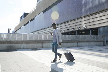 Männlicher Unternehmer beim Ziehen des Gepäcks und Benutzen des Mobiltelefons am Bahnhof in der Stadt - PMF01551