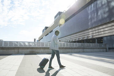 Male professional pulling luggage while using mobile phone against station in city - PMF01552