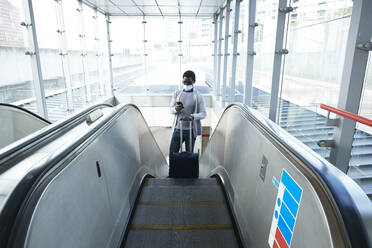 Businessman with luggage using smart phone while standing on escalator at railroad station during COVID-19 - PMF01567
