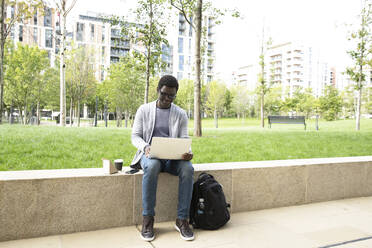 Geschäftsmann, der einen Laptop benutzt, während er beim Mittagessen auf einer Stützmauer in der Stadt sitzt - PMF01576