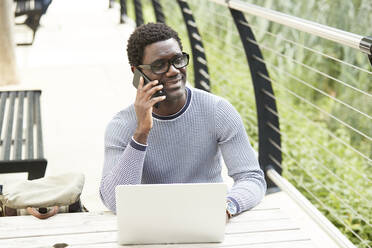 Male professional talking on mobile phone looking away while sitting with laptop at table - PMF01603