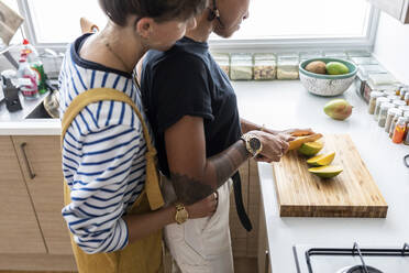 Freundin schneidet Obst, während die Frau sie von hinten in der Küche zu Hause umarmt - WPEF03584