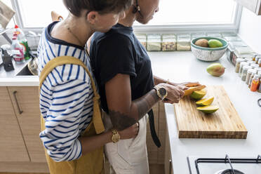 Girlfriend cutting fruit while woman embracing her from behind in kitchen at home - WPEF03584