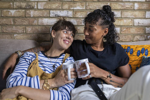 Lächelnde lesbische Paar Toasting Teetasse beim Sitzen auf dem Sofa zu Hause - WPEF03598