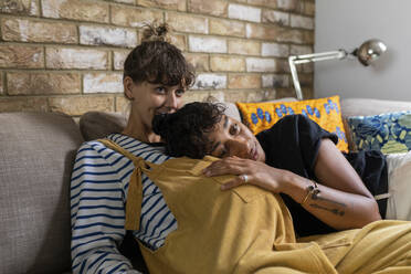 Woman leaning on pregnant girlfriend while sitting on sofa at home - WPEF03600