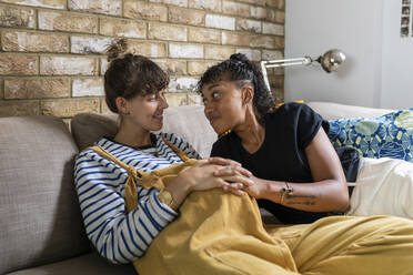 Smiling lesbian couple sitting with hands clasped on sofa at home - WPEF03601