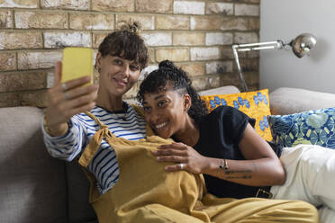 Pregnant woman taking selfie on mobile phone with girlfriend while sitting on sofa at home - WPEF03607