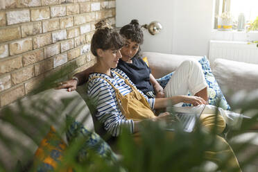 Freundin betrachtet Frau, die einen Laptop benutzt, während sie zu Hause auf dem Sofa sitzt - WPEF03608