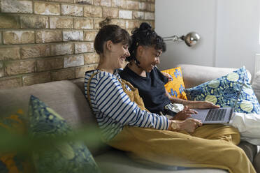 Lesbisches Paar, das lächelnd einen Laptop benutzt und zu Hause auf dem Sofa sitzt - WPEF03615