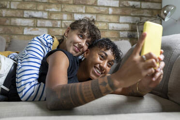 Smiling woman taking selfie on mobile phone with girlfriend while lying on sofa at home - WPEF03628