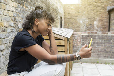 Lächelnde Frau, die ein Mobiltelefon benutzt, während sie an einer Mauer im Hinterhof sitzt - WPEF03635