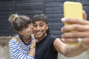 Lächelnde Frau, die ein Selfie auf dem Handy macht, während ihre Freundin sie küsst und im Hinterhof steht - WPEF03644