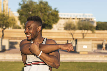 Smiling man stretching arms while standing at park - PNAF00189