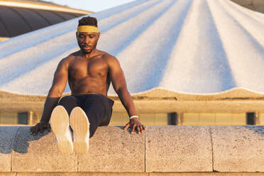 Athlete doing abdominal muscle exercise while sitting on retaining wall at park - PNAF00200