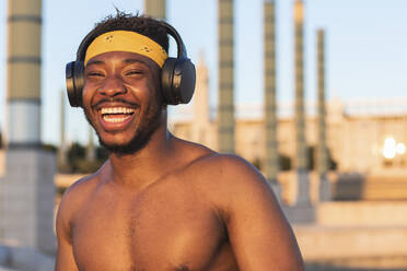 Shirtless man wearing headphones laughing while standing at park - PNAF00201