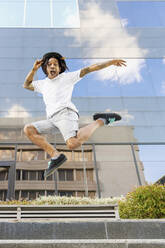 Young man wearing hat screaming while jumping against modern building in city - IFRF00086