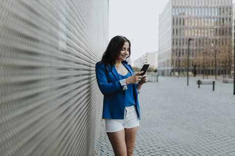 Smiling businesswoman using smart phone while standing on footpath in city stock photo