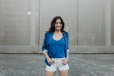 Smiling woman standing on footpath against wall - GMLF00822
