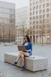 Happy businesswoman using laptop while sitting on bench in city - GMLF00826