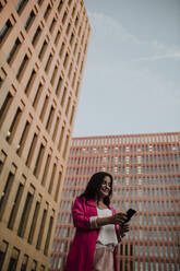 Weibliche Unternehmerin schreibt Textnachrichten auf ihrem Smartphone, während sie vor einem Gebäude steht - GMLF00841