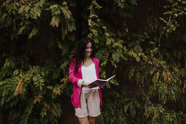 Female entrepreneur reading diary while standing against tree - GMLF00842