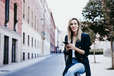 Lächelnde modische Frau, die wegschaut, während sie ein Mobiltelefon in der Stadt hält - EBBF01549