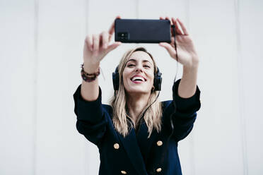 Lächelnde Frau, die ein Selfie mit ihrem Smartphone macht, während sie an der Wand Musik hört - EBBF01554