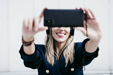 Smiling blond woman taking selfie while listening music against wall - EBBF01555