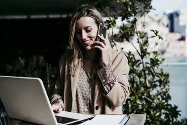 Lächelnde Geschäftsfrau, die einen Laptop benutzt, während sie mit einem Mobiltelefon im Büro spricht - EBBF01559
