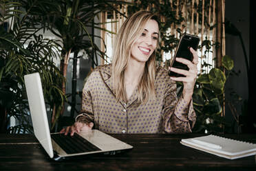 Lächelnde blonde Geschäftsfrau, die ein Mobiltelefon benutzt, während sie mit einem Laptop in einem kreativen Büro sitzt - EBBF01567