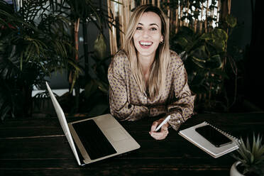 Happy blond female entrepreneur sitting with laptop at desk in office - EBBF01568