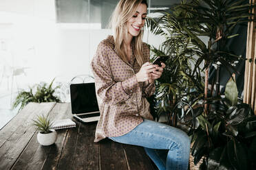 Blond businesswoman using mobile phone while sitting on desk in office - EBBF01570