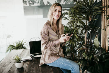 Lächelnde schöne weibliche professionelle hält Smartphone beim Sitzen auf dem Schreibtisch im Büro - EBBF01571