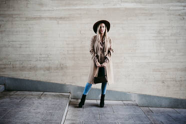 Stylish blond woman wearing coat and hat standing against wall - EBBF01574