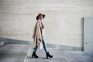 Stilvolle Frau, die einen Mantel trägt, während sie auf einer Treppe an der Wand geht - EBBF01577