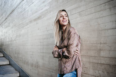 Happy beautiful woman with arms crossed looking away while leaning on wall - EBBF01580