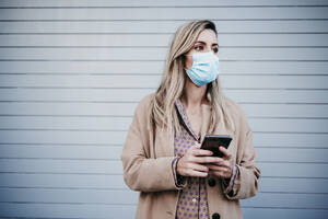 Beautiful blond woman wearing protective face mask while holding mobile phone against wall - EBBF01582