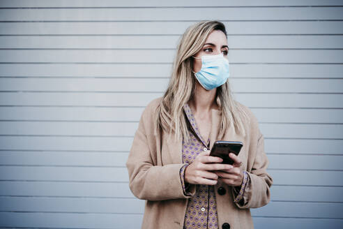 Beautiful blond woman wearing protective face mask while holding mobile phone against wall - EBBF01582