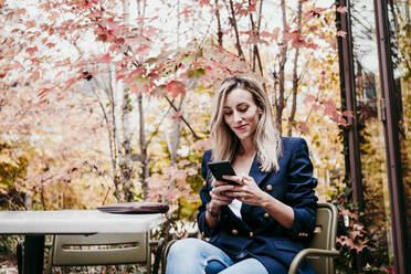 Blonde Frau im mittleren Erwachsenenalter, die ein Smartphone benutzt, während sie auf der Terrasse vor den Herbstbäumen sitzt - EBBF01585