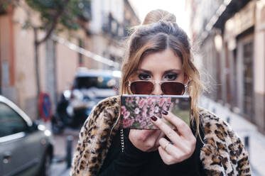 Junge Frau benutzt ein Mobiltelefon, um Lippenstift aufzutragen, während sie auf der Straße steht - JMPF00618